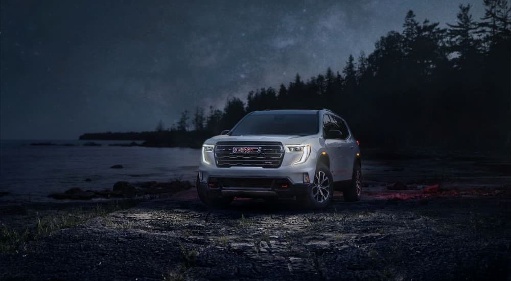 A GMC Acadia for sale, a white 2024 GMC Acadia AT4, is shown parked on rocks with the headlights illuminated.