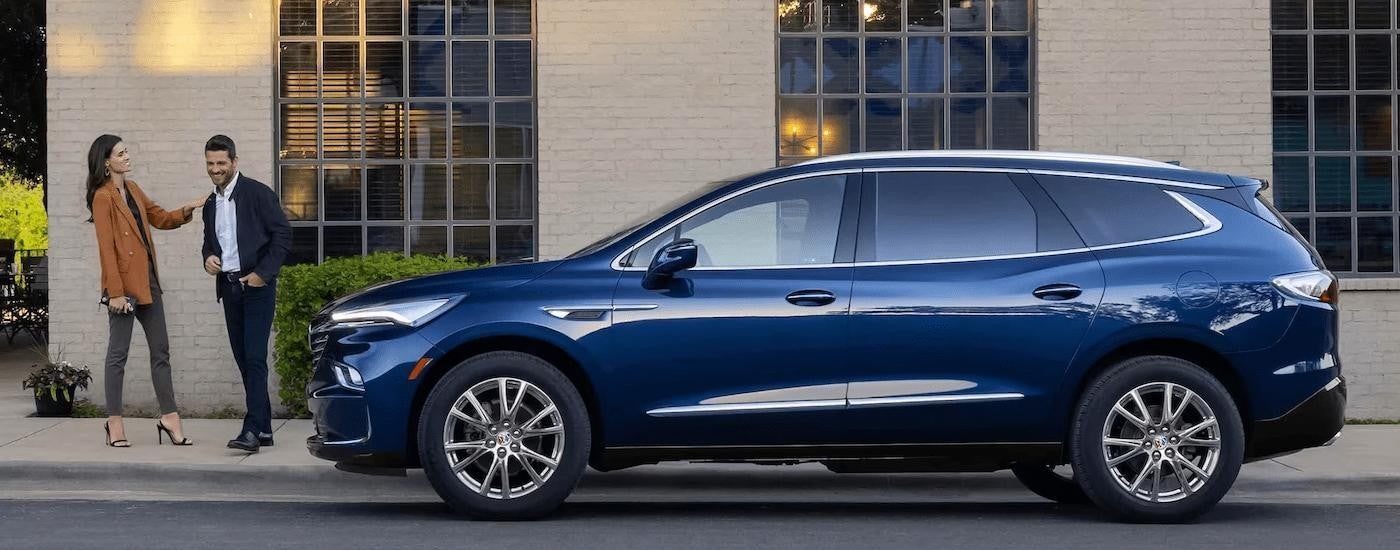 A well-dressed couple laughing beside a blue 2023 Buick Enclave parked on a city street.