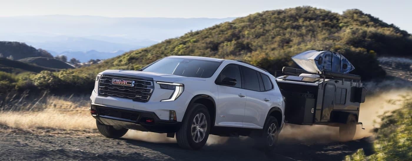 A white 2025 GMC Acadia AT4 is towing a small camper trailer on a dusty gravel road.